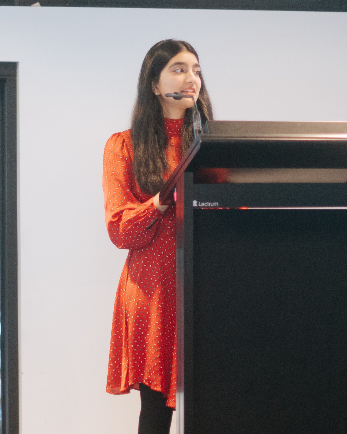 Anya Gupta in a red patterned dress speaking into a headset microphone at a lectern in front of a white wall, at the Melbourne Diwali MD25 launch.