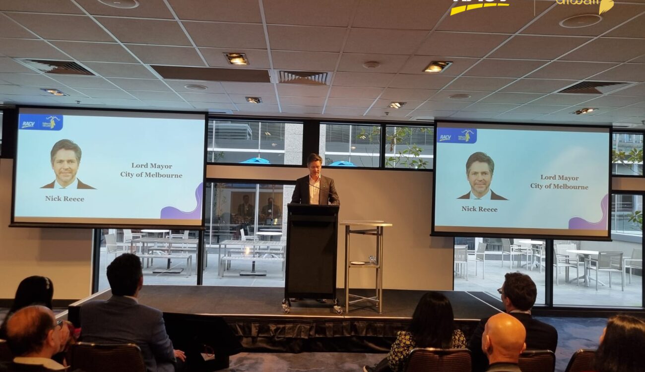 A speaker at a lectern between two screens showing a portrait of Nick Reece, Lord Mayor of Melbourne, addressing a seated audience at the 2024 Melbourne Diwali launch.