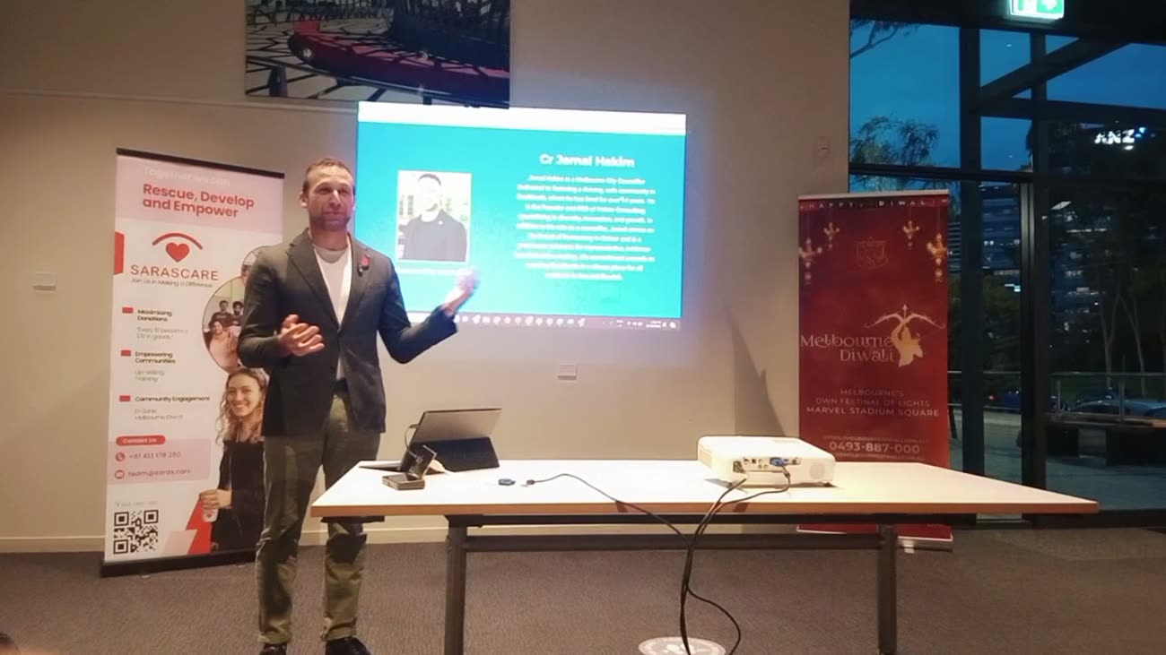 Cr Jamal Hakim speaking at the D-Zone launch in a Docklands community room, between a Saras.Care banner and a Melbourne Diwali banner, with his title slide projected behind him.
