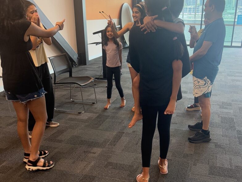 A group of teenagers standing in a loose circle mid drama exercise in a carpeted room with an orange wall and harbour windows.