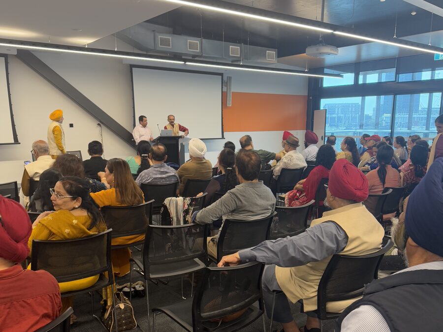 A room of seated older people, many in turbans and shawls, facing two speakers at a lectern.