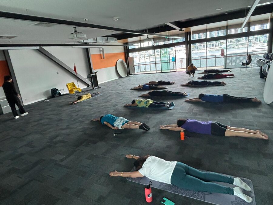 About a dozen people lying on mats in savasana on the floor of a large community room with harbour windows.