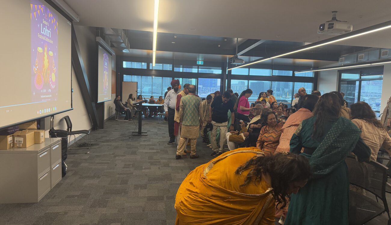 A community room with floor-to-ceiling windows onto the harbour, full of people standing and seated in front of a screen reading Lohri.
