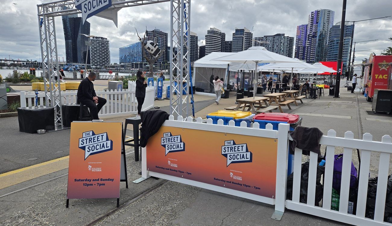 The Street Social on Harbour Esplanade: a truss archway, marquees, picnic tables and food trucks with the Docklands towers behind.